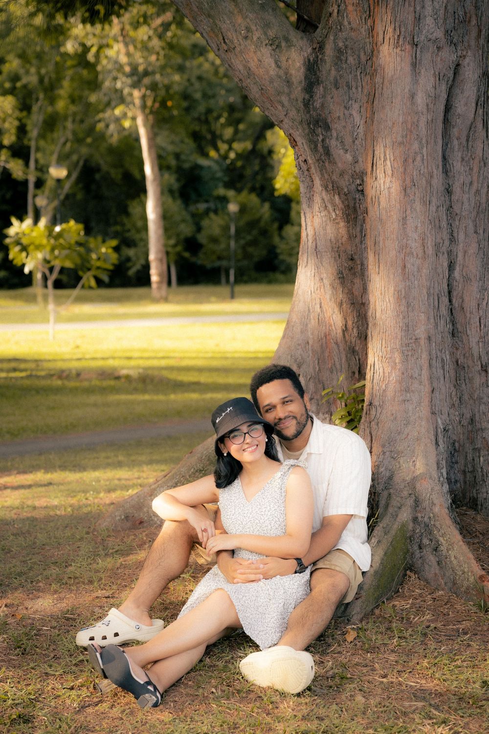 A couple relaxes together under large trees in a park during golden hour.