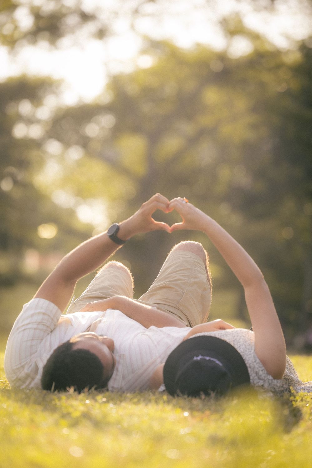 Couple in white outfits lying on grass while making heart shapes with their hands at sunset in a park setting.