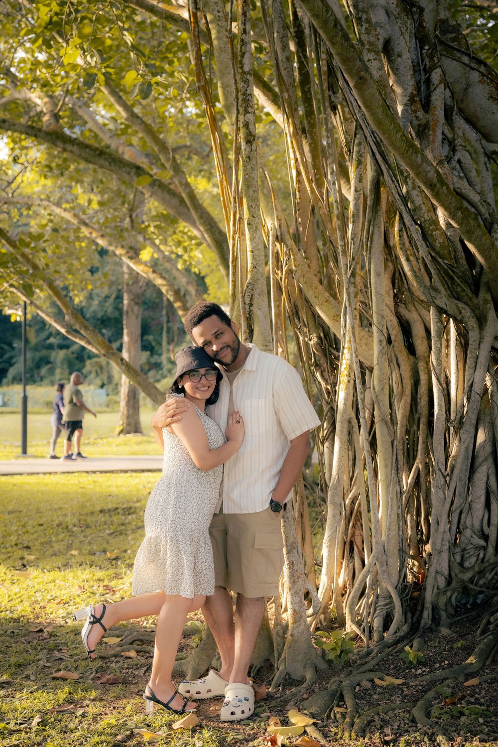 A couple embraces while standing beneath large banyan trees in a sunlit park during golden hour.
