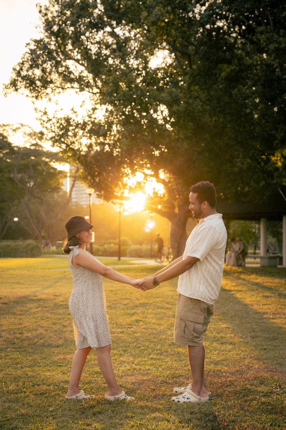 A couple holds hands in a park during golden hour sunset as sun rays shine through trees on a summer evening.