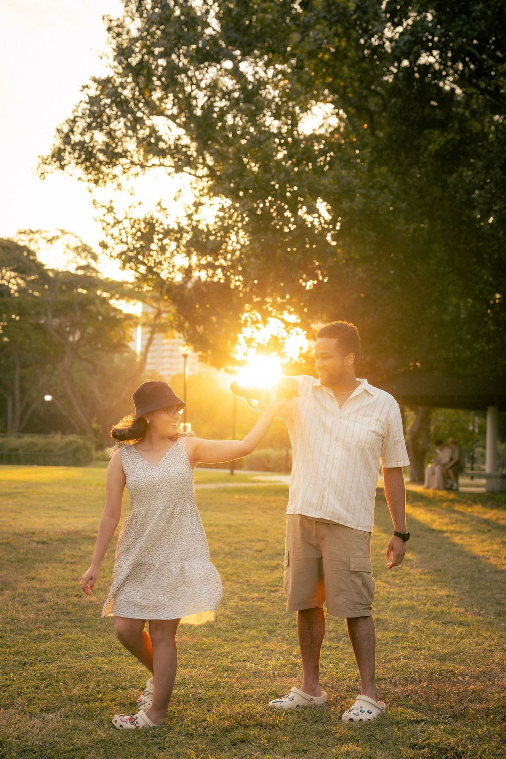 Two people in white outfits stand together on grassy field during golden hour sunset creating a romantic silhouette.