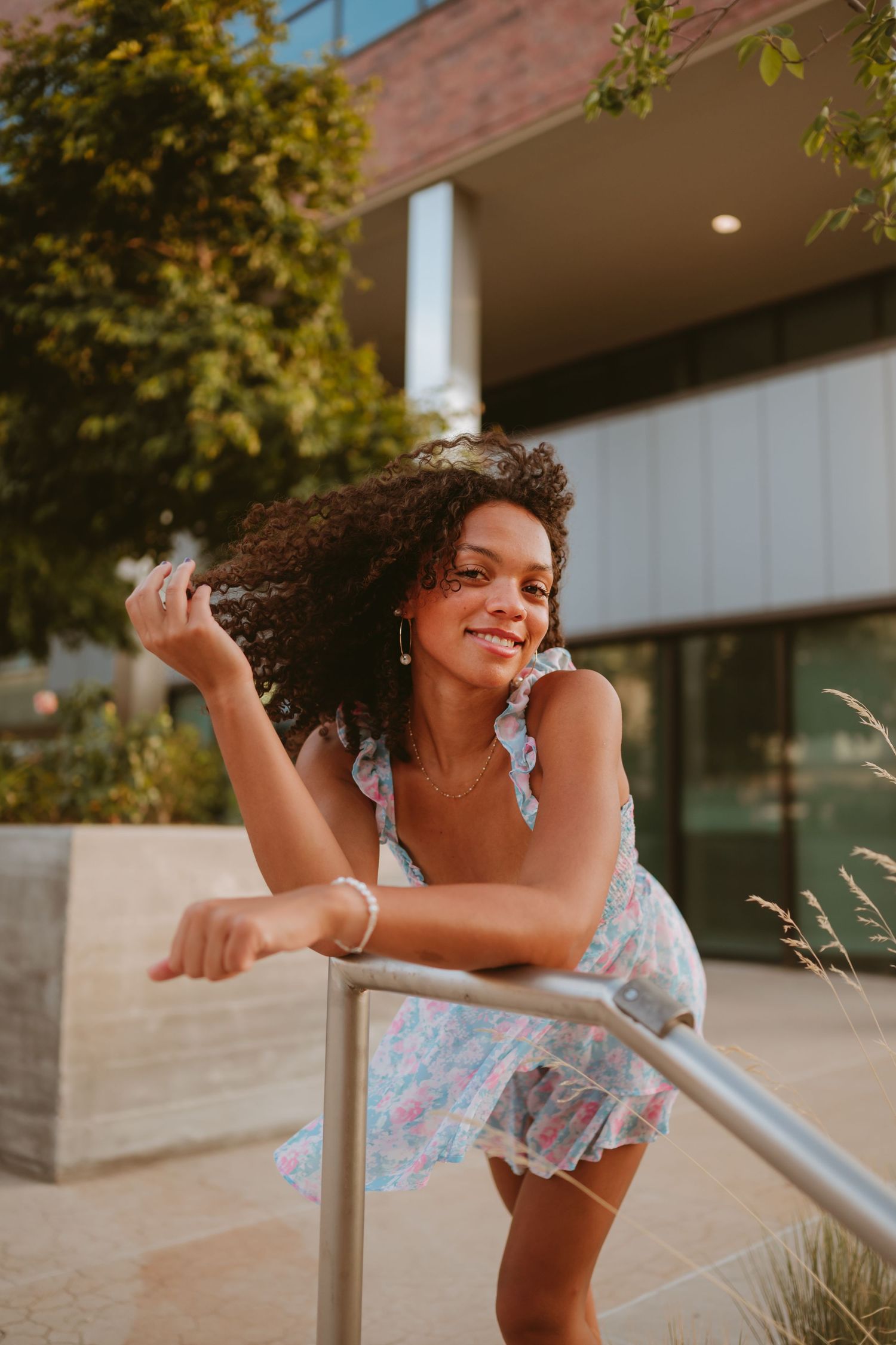 Young woman in floral dress leans against railing with curly hair blowing in wind on warm sunny day.