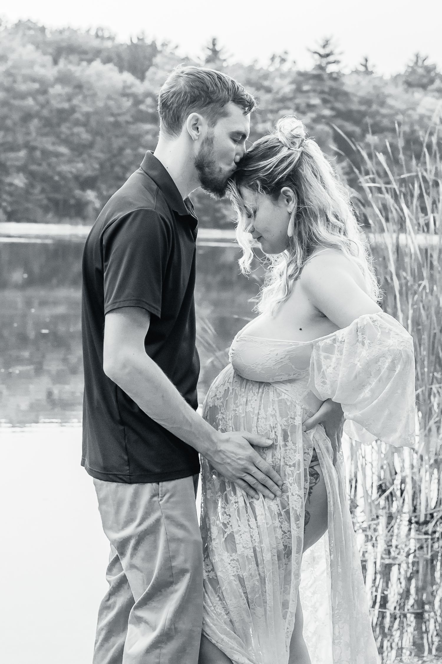 Romantic black and white maternity photo by a lake with tall reeds and trees in background.