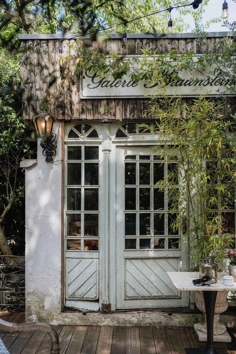 Rustic storefront with blue-gray trim windows and doors surrounded by lush greenery and vines.