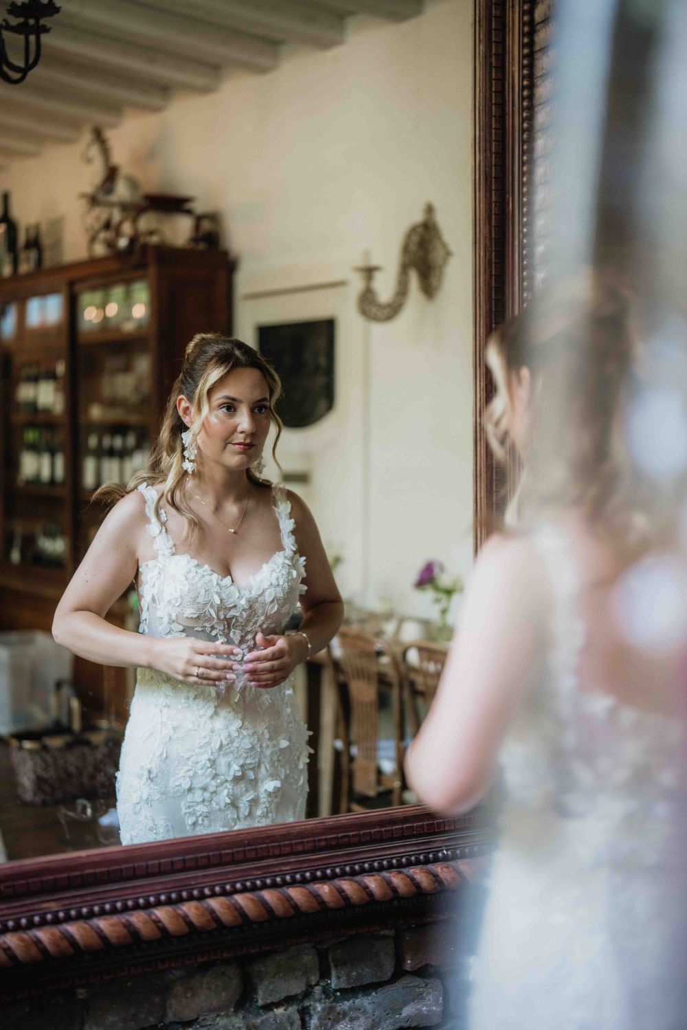 Elegant bride in lace dress looking at her reflection in antique wooden framed mirror.