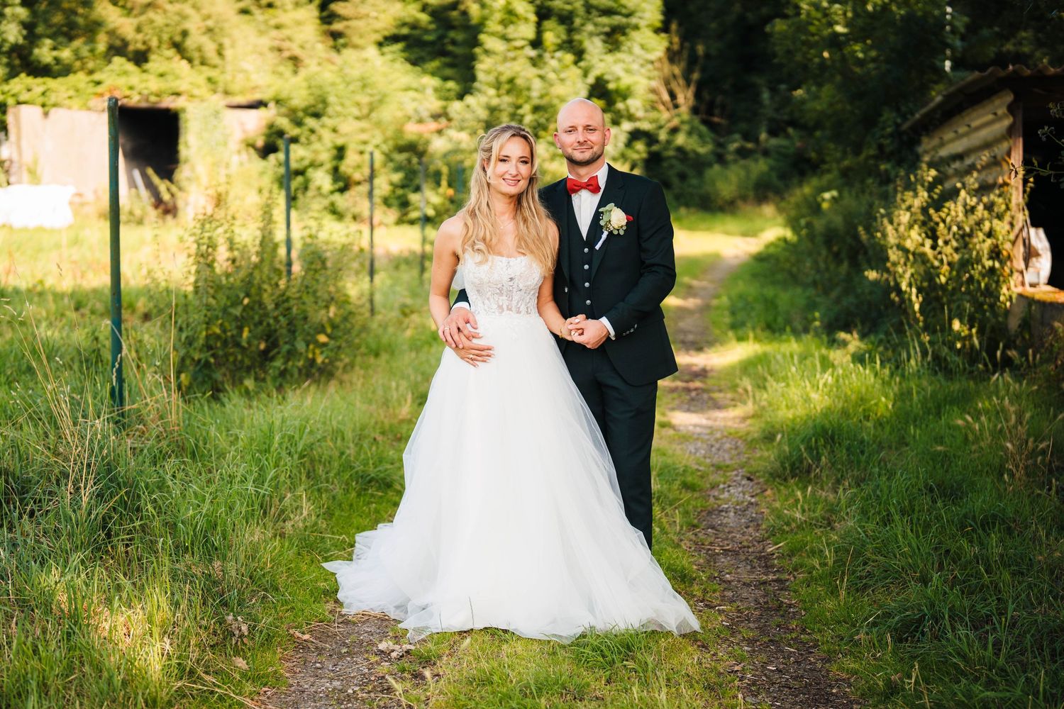 A couple walks along a sunlit path through green summer fields on their wedding day.