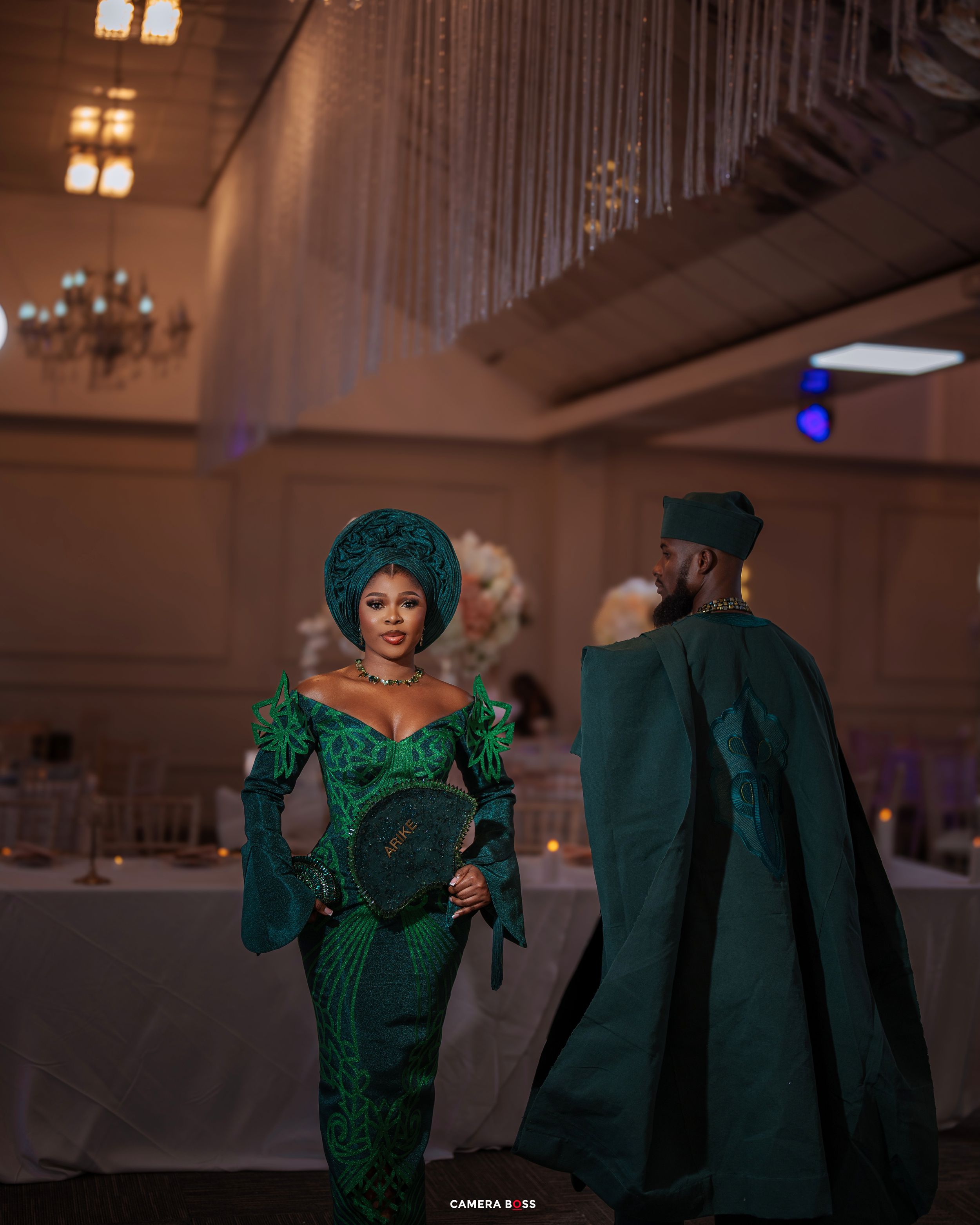 Two people in ornate emerald green traditional attire with matching headpieces at an elegant indoor venue.