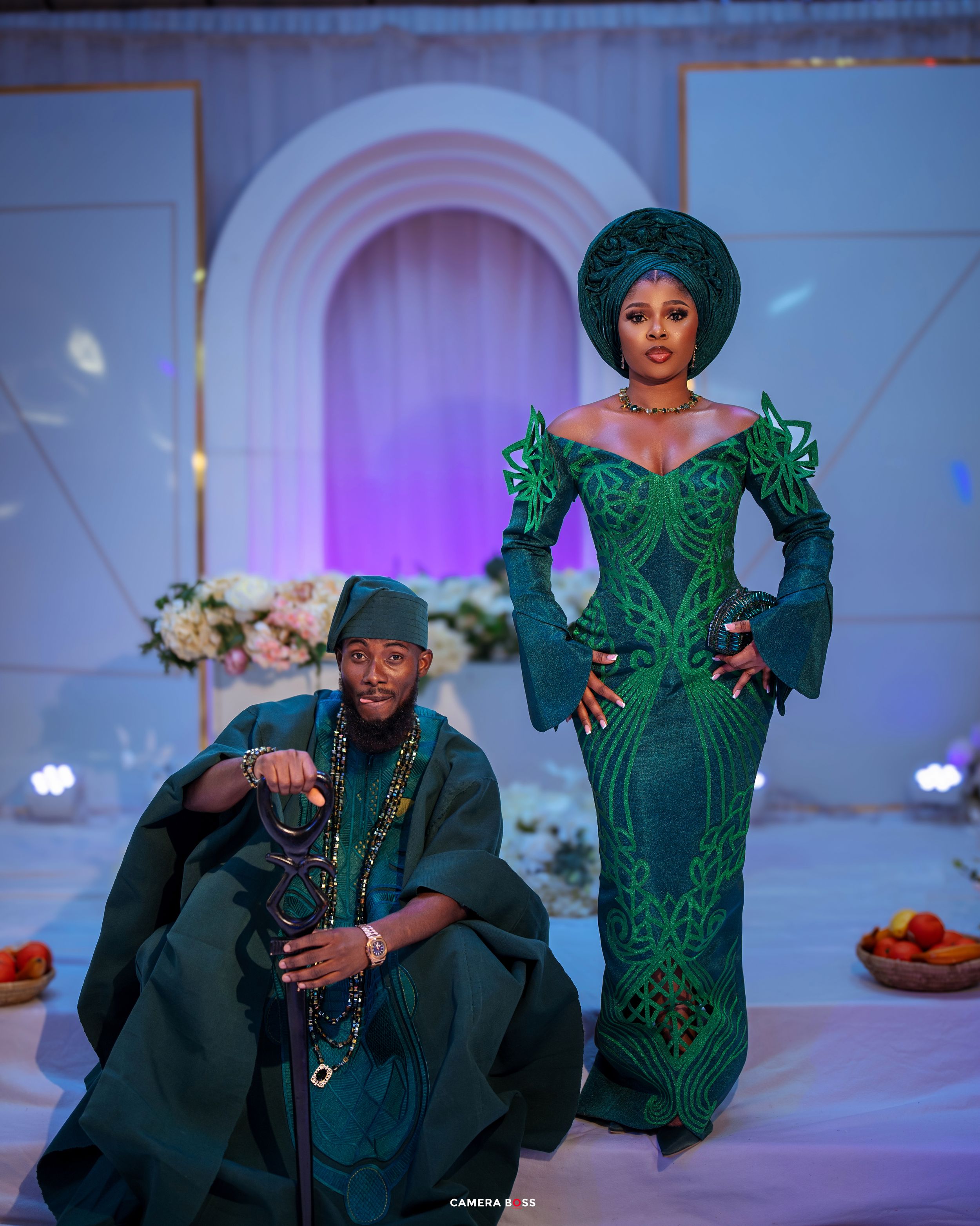Two people in elegant emerald green traditional African attire pose together in front of an archway with purple lighting effects.