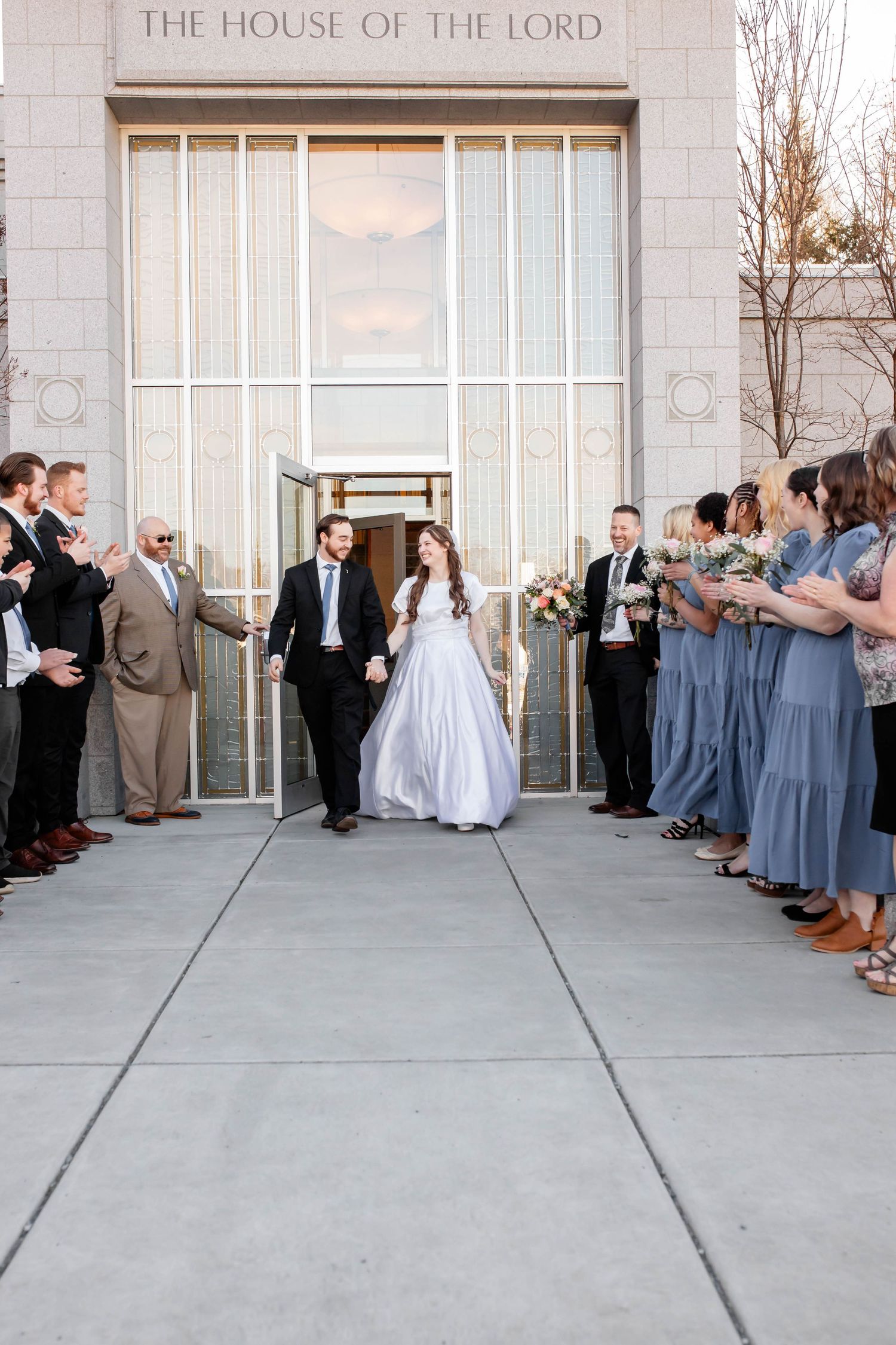 A wedding party exits a House of the Lord temple with bridesmaids in blue dresses and groomsmen lining the walkway.