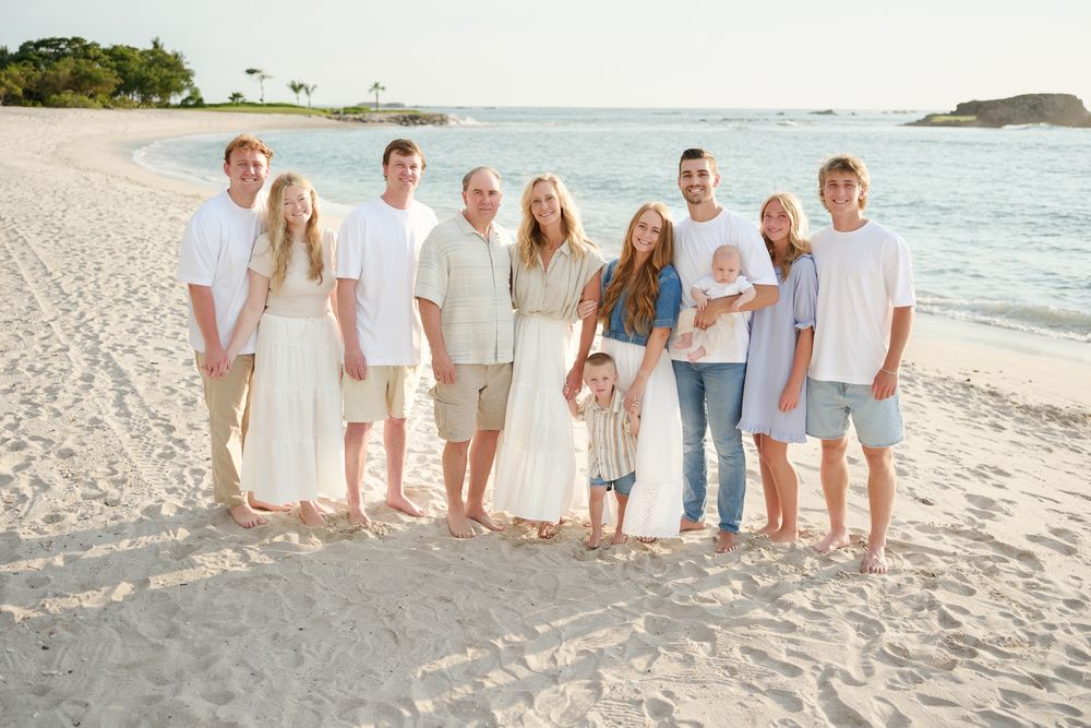 A group wearing light beige and white outfits stands together on a tropical beach with turquoise water and islands behind them.