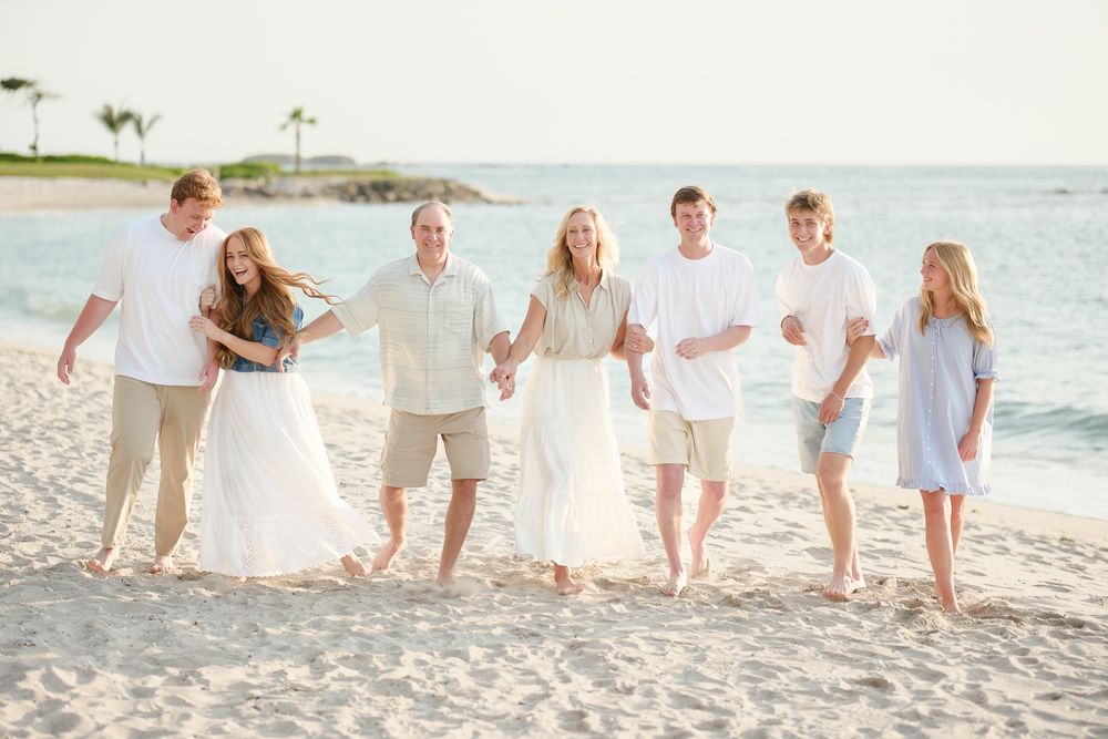 A group of people in white and light colored clothing walk together joyfully along a tropical beach at sunset.
