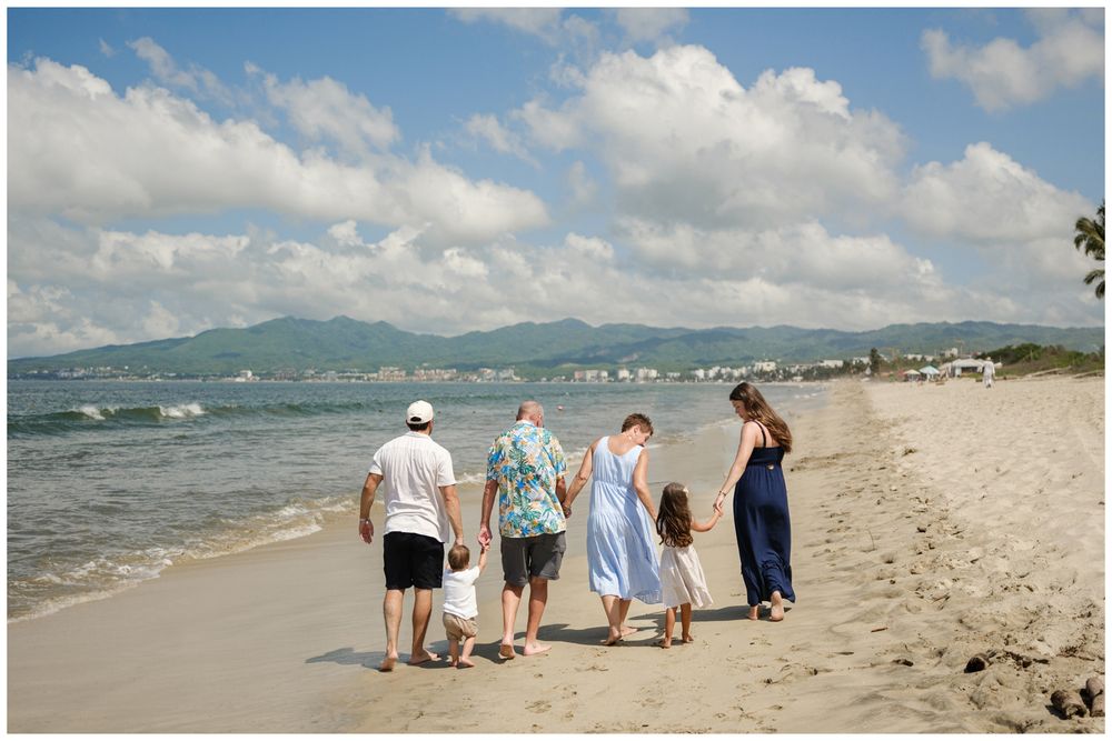 Group of people walking along a scenic tropical beach with mountains visible in the distance and cloudy skies overhead.