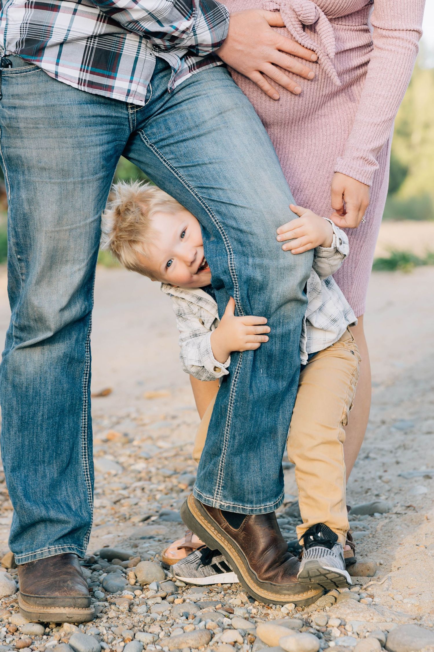 Toddler playfully peeking through parents legs on a rocky outdoor path.