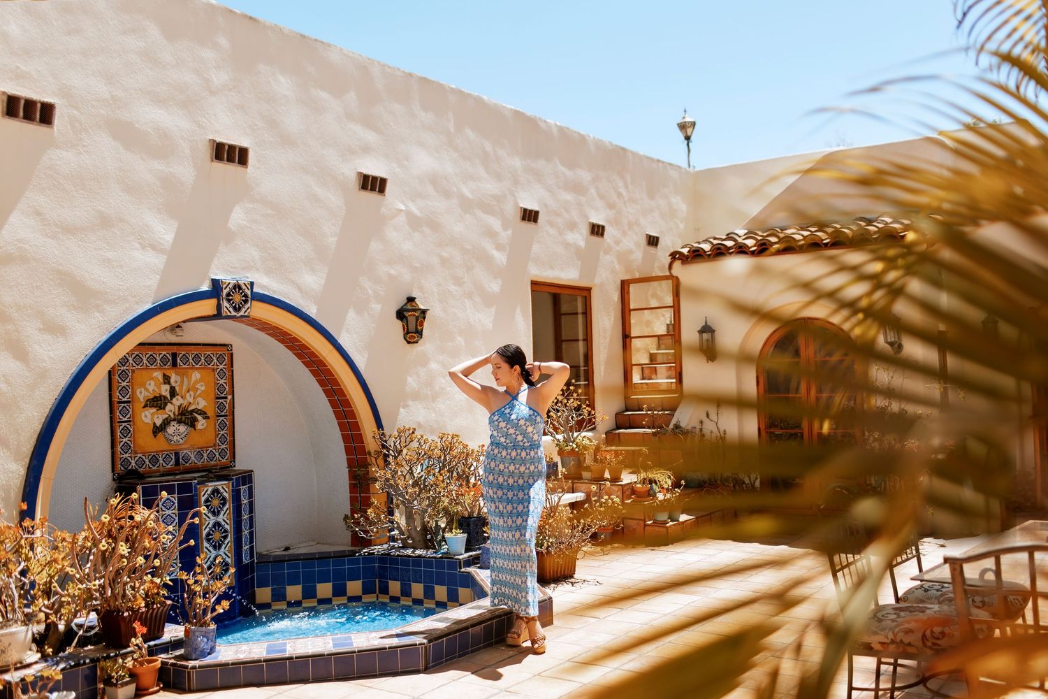 A woman poses in front of a Spanish style home in a blue dress surrounded by succulent plants.