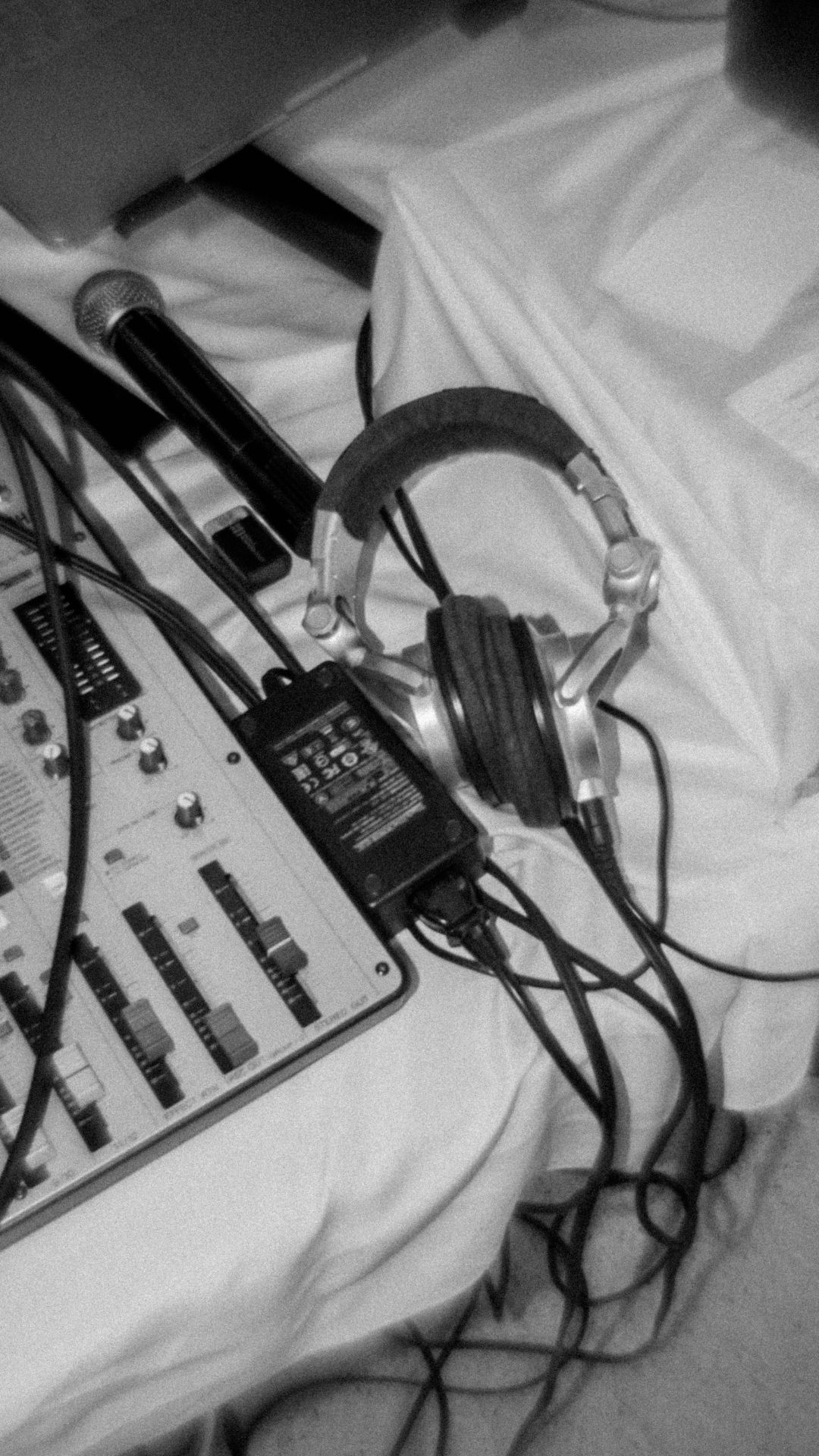 Black and white photo of audio equipment including headphones, microphone, and mixing board on white fabric.