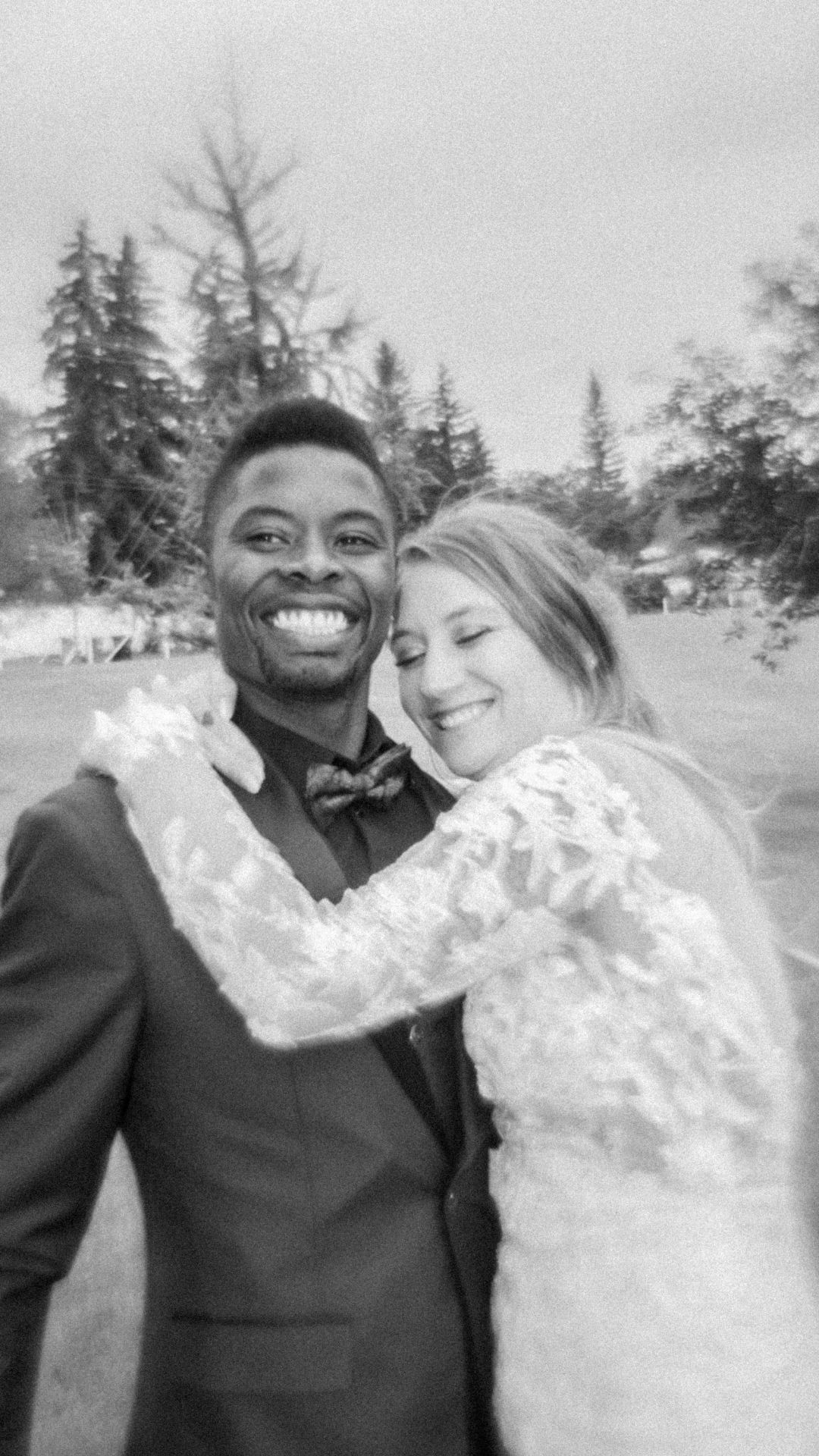 A joyful black and white wedding photo showing a couple embracing outdoors with evergreen trees in the background.