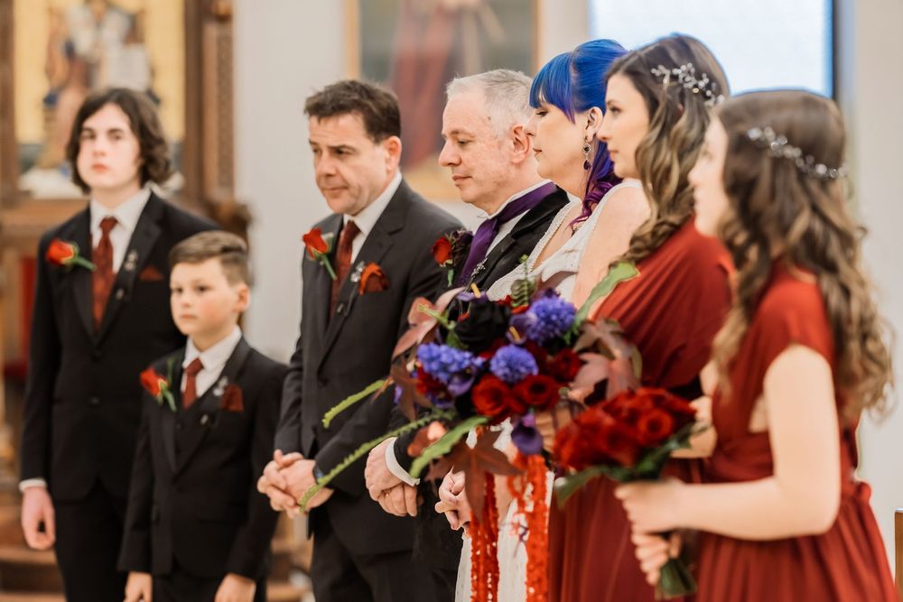 Wedding party members in red and black attire stand in line at church ceremony with blue and purple bouquets.