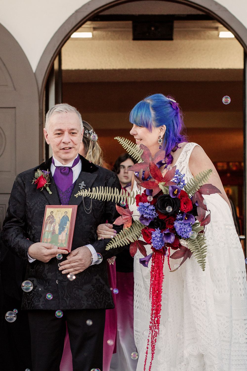 Wedding couple stands in archway, bride wears white dress with blue hair, holding colorful bouquet with purple and red flowers.