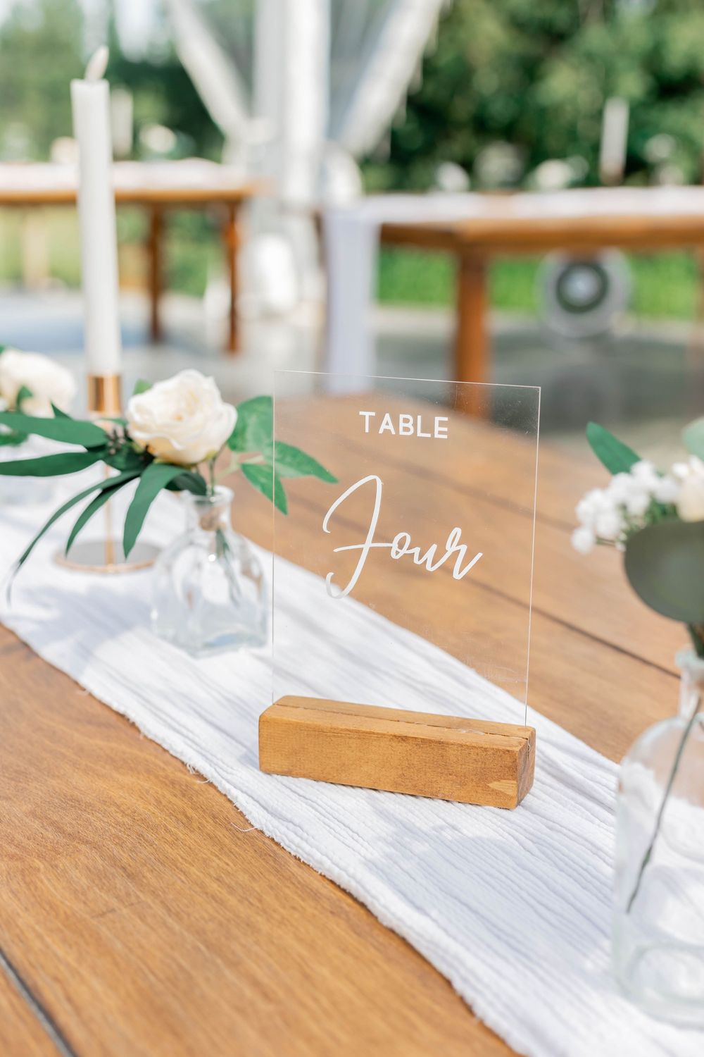 Clear acrylic wedding table number sign reading 'Table Four' with white flowers and wooden base on rustic table decor.