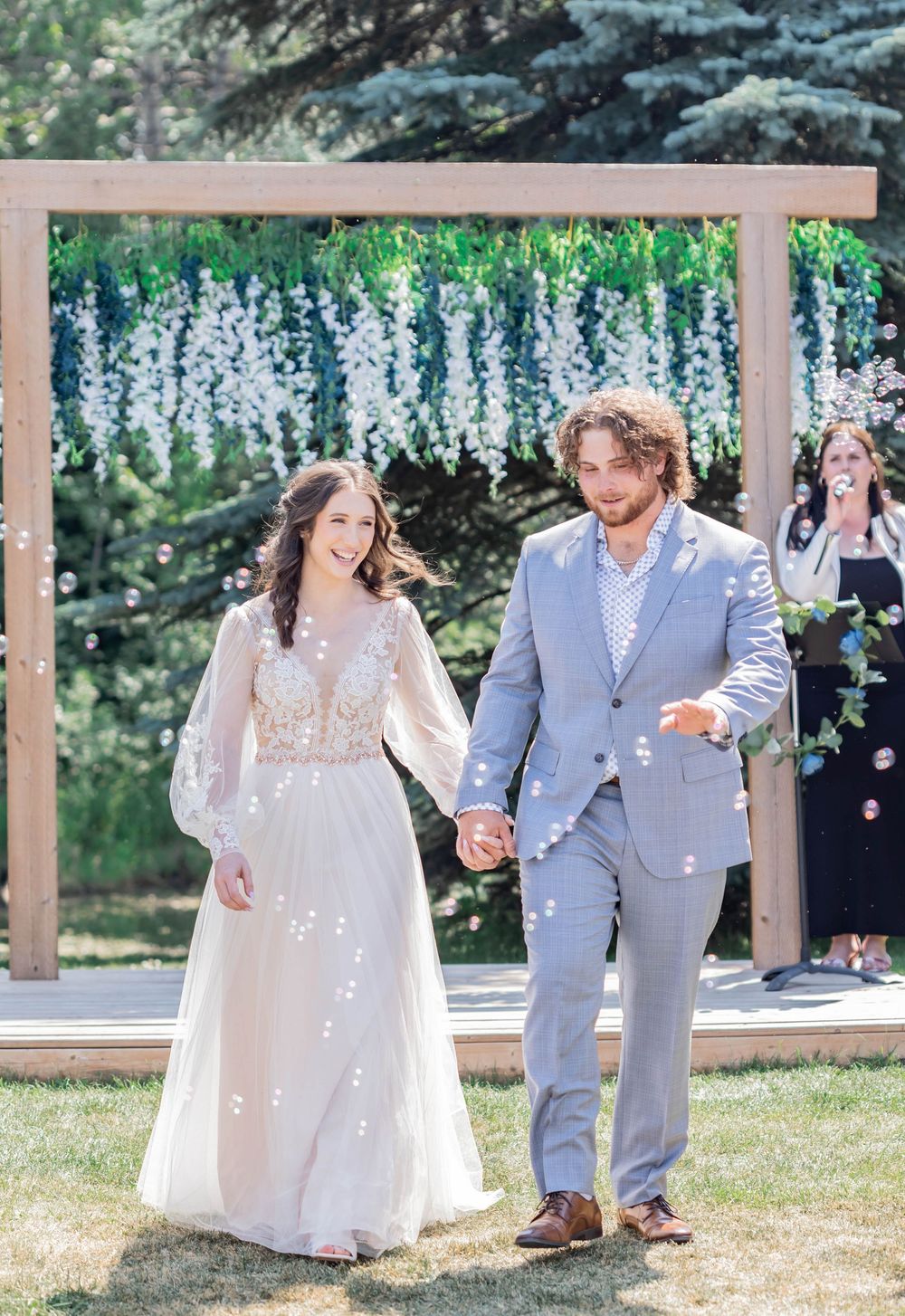 Floral wedding ceremony under white wisteria arch with couple in light blue suit and flowing lace gown walking together.