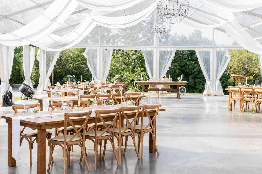 Long wooden banquet tables with crossback chairs are arranged under an elegant white tent for an outdoor wedding reception.