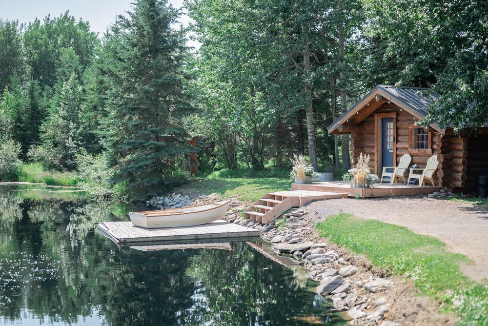 A rustic wooden cabin sits on the shore of a serene lake with docked boats and surrounded by evergreen trees.