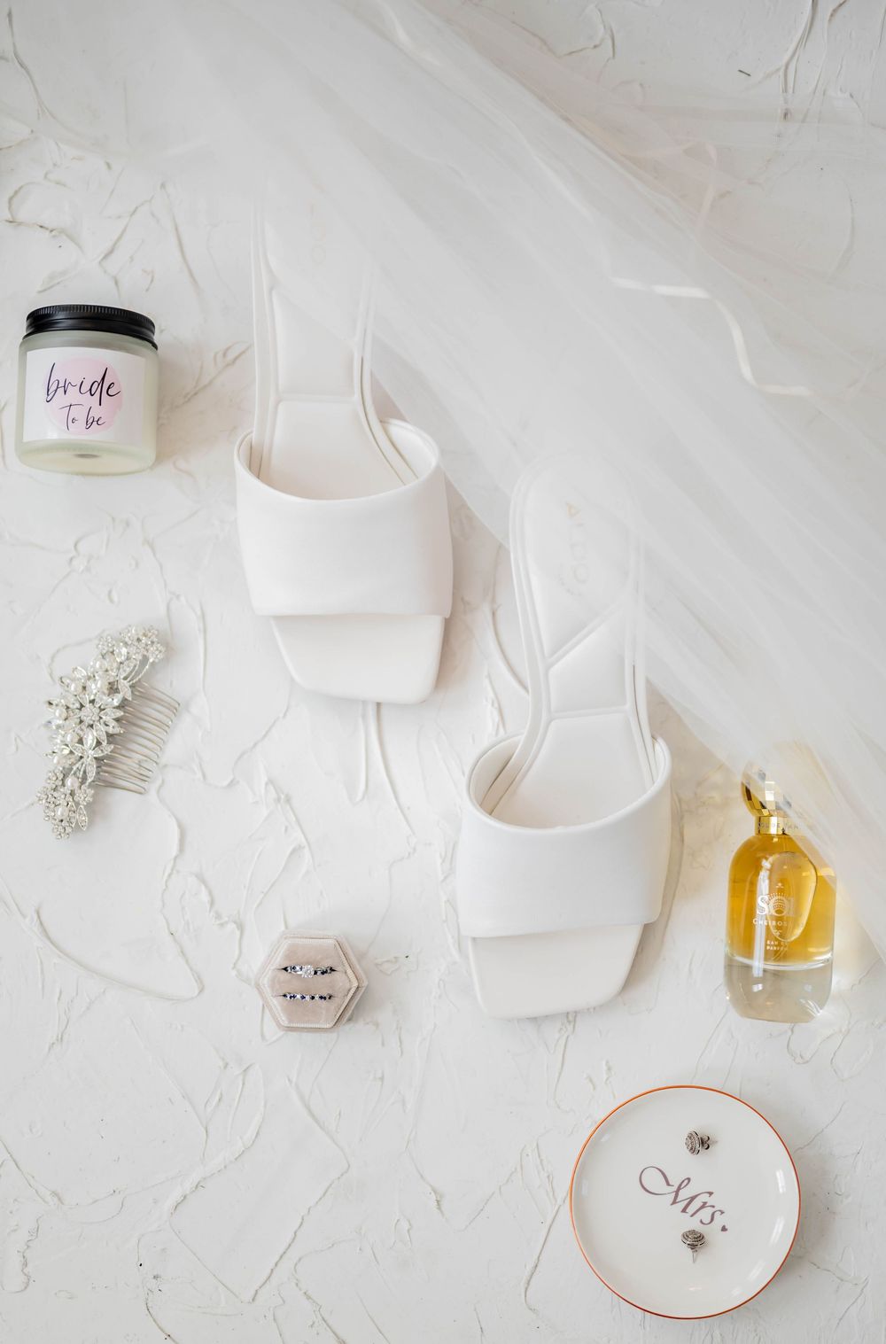 Wedding details including perfume bottles, heart shapes, and delicate bridal accessories arranged on white textured backdrop.