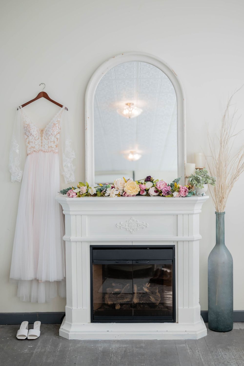 Elegant white wedding dress hangs beside a decorated fireplace mantel with fresh flowers and pampas grass in a vase.