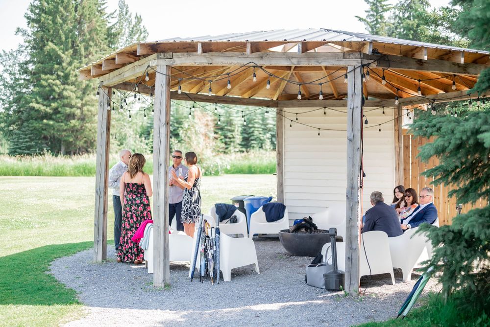 People gather under a rustic wooden pergola with string lights on a grassy lawn near evergreen trees.