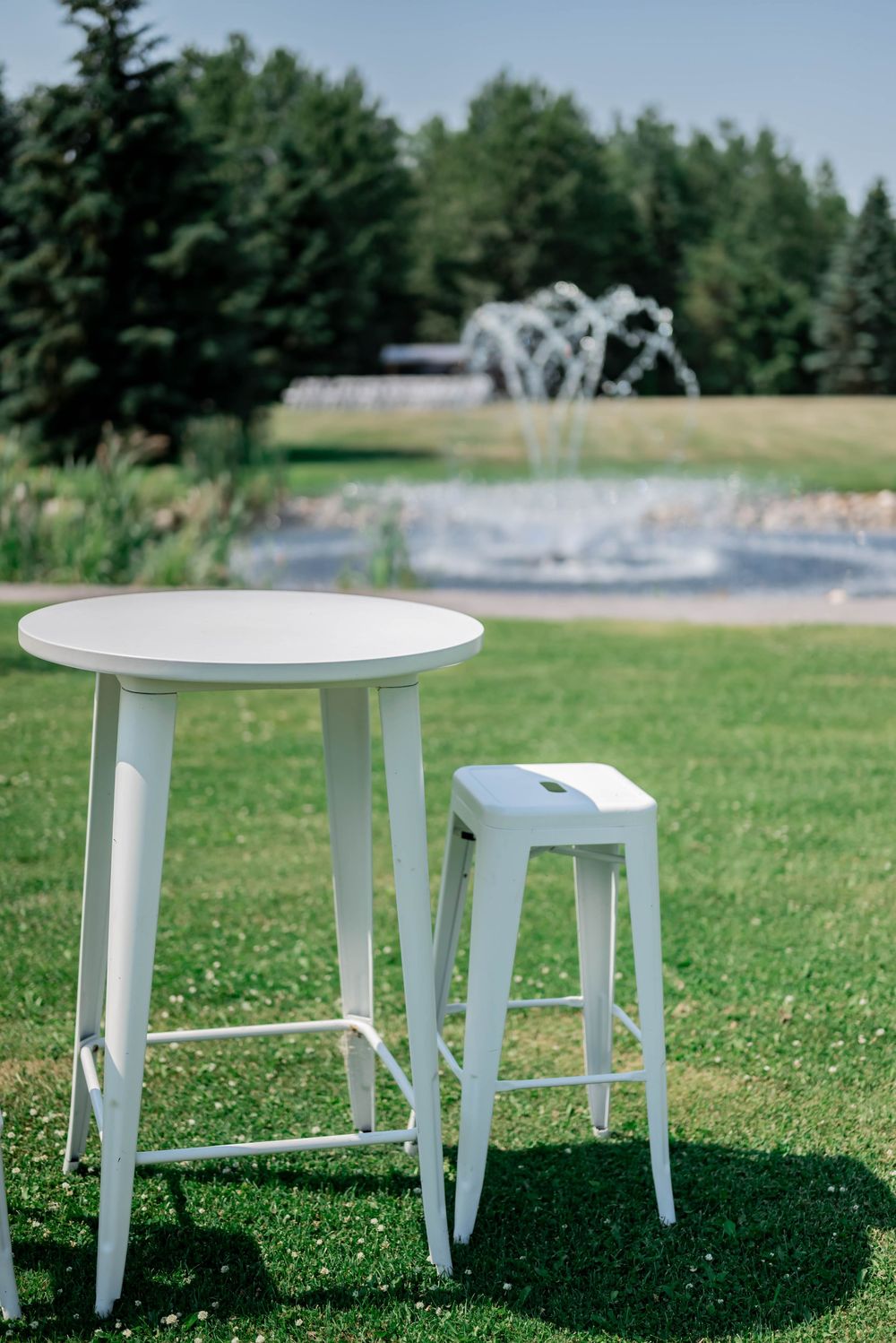 Four white round side tables arranged on a sunny green lawn with trees and a fountain in the background.