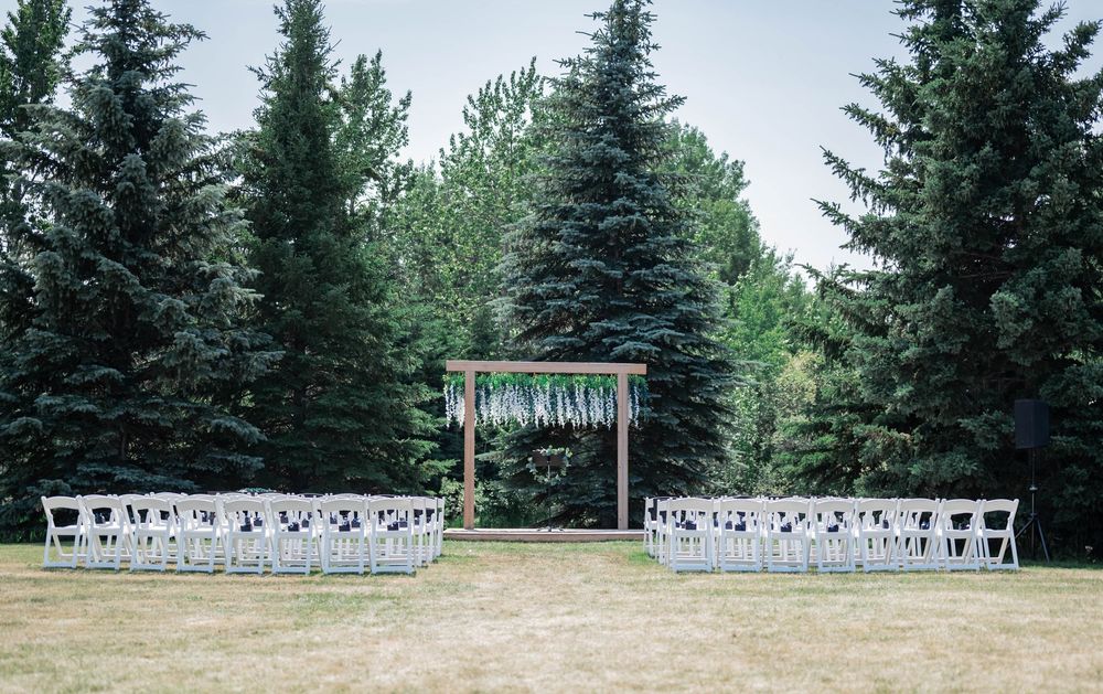 White chairs and a wooden arch decorated with florals create an intimate outdoor wedding ceremony setup among evergreen trees.