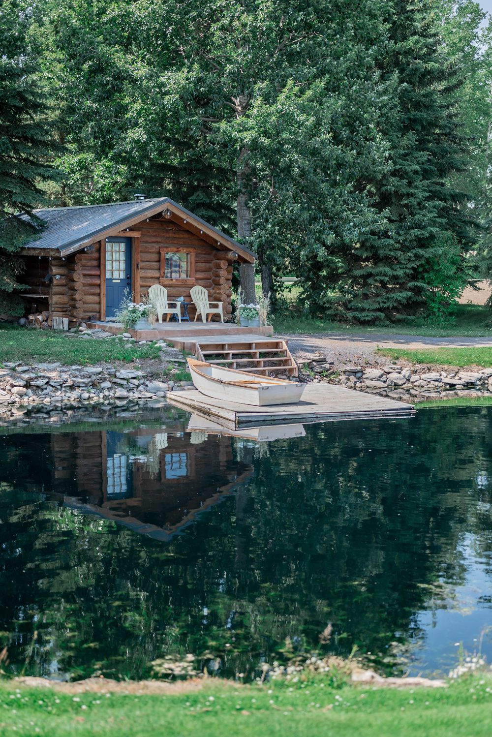 A rustic log cabin with a wooden dock sits beside a reflective pond surrounded by evergreen trees.
