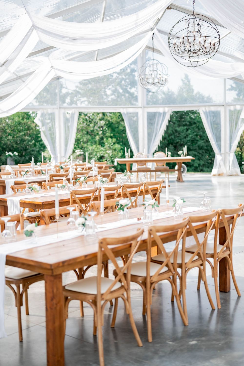 Elegant wedding reception setup with rustic farm tables and crossback chairs under white draped tent with crystal chandeliers.