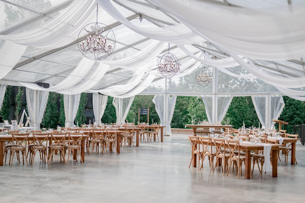 Rows of elegant wooden cross-back chairs arranged under white draped tent canopies for an outdoor wedding reception.