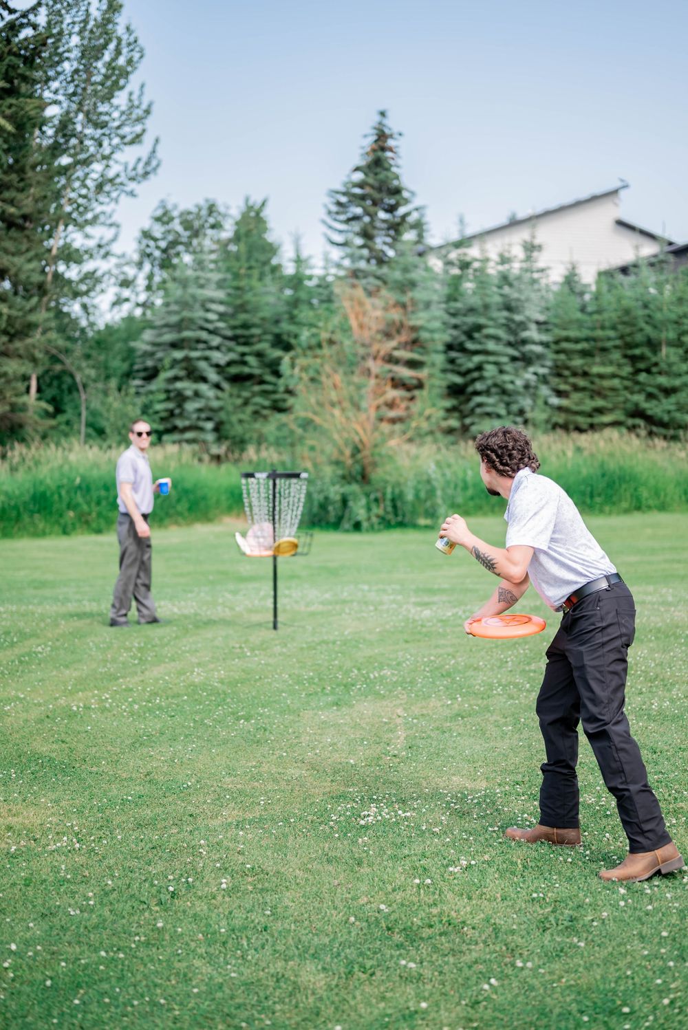 Two people playing disc golf on a grassy course with evergreen trees and a building in the background.