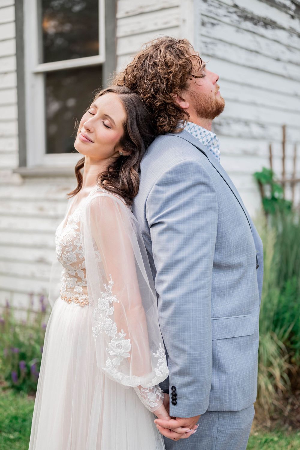 Romantic couple in grey suit and lace dress share intimate moments against white farmhouse wall at outdoor wedding.