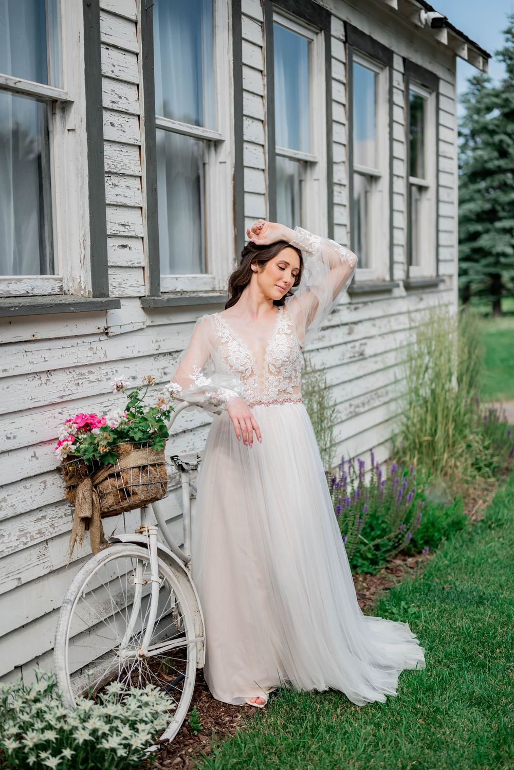 A vintage white bicycle adorned with flowers stands against a rustic building as a woman in a flowing white dress poses gracefully.