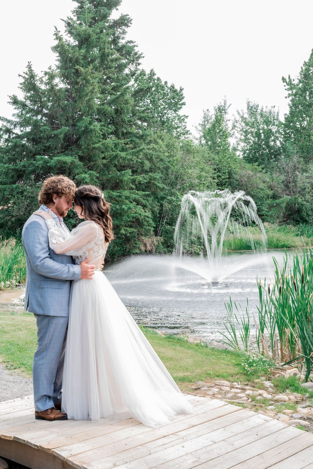 A couple embraces on a wooden deck overlooking a pond with a fountain in a serene garden setting.