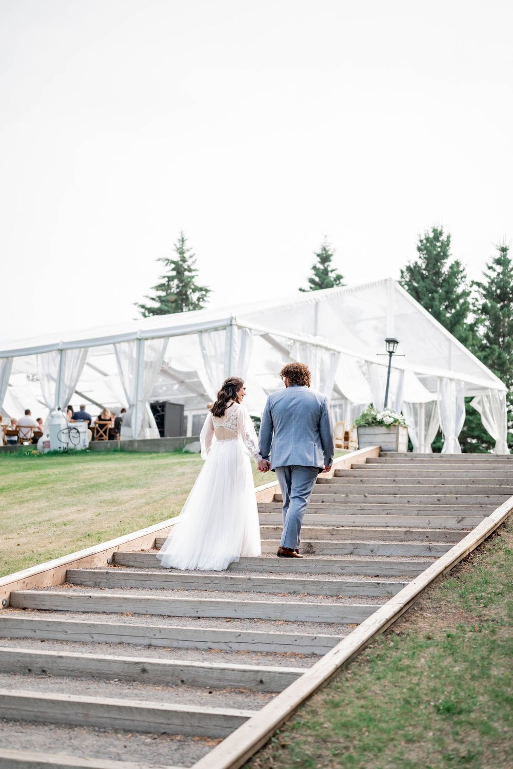 A couple ascending wide wooden stairs outdoors with a white tent structure visible at the top.