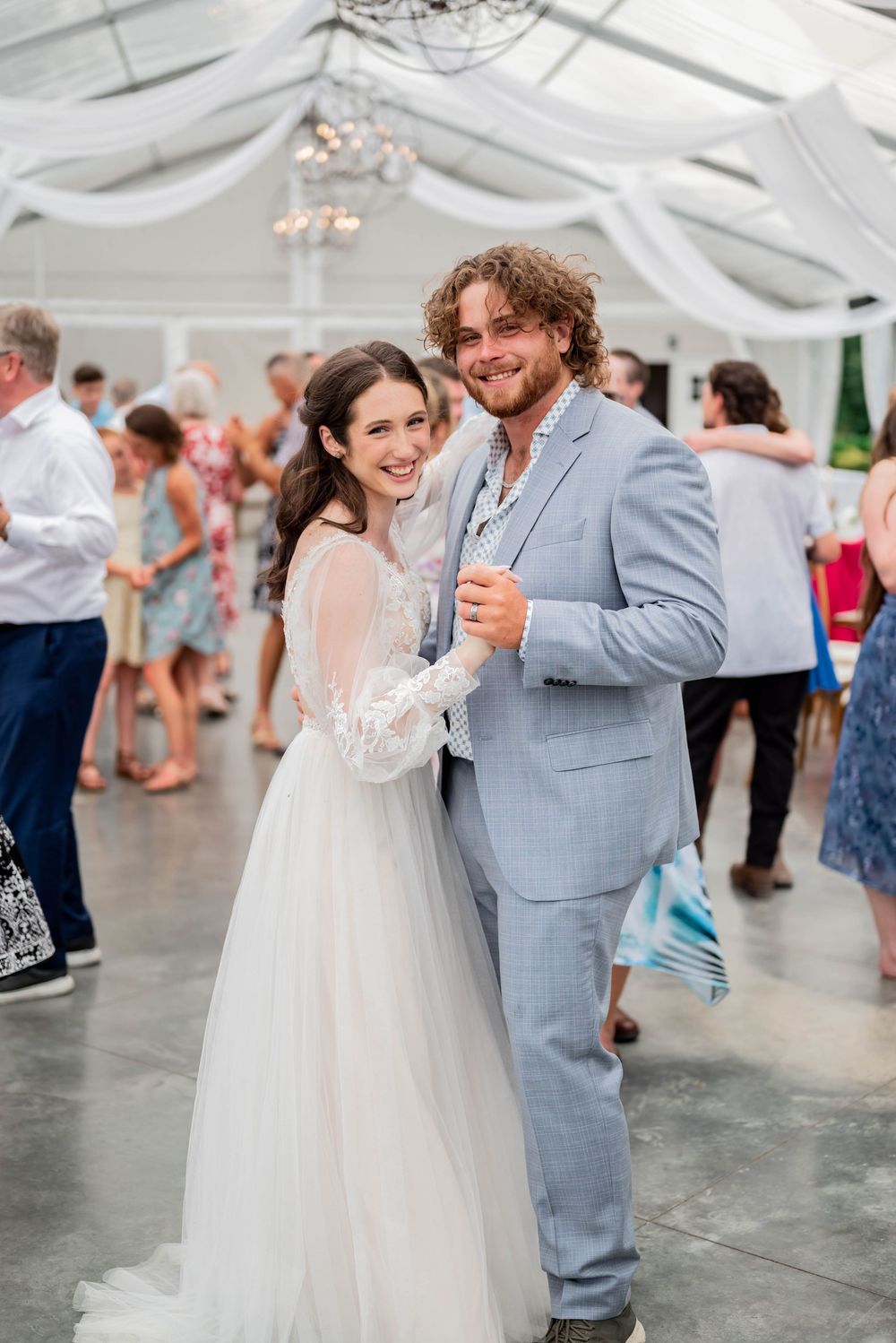 Happy couple dances together at a wedding reception under a white tent with decorative draping and lights.