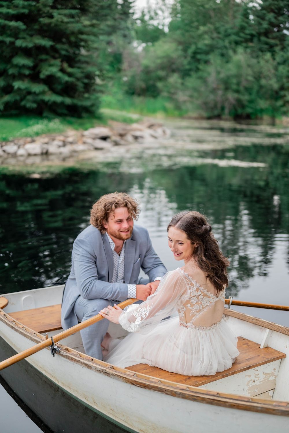 A couple shares a romantic moment in a wooden rowboat on a serene lake surrounded by evergreen trees.