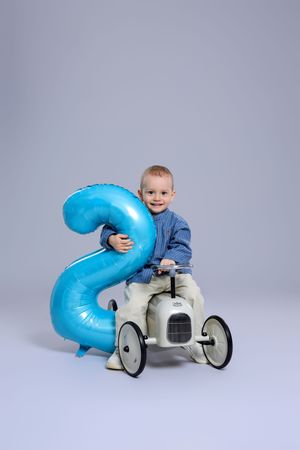 Smiling toddler boy sitting on vintage toy car holding blue number 2 balloon on gray background.