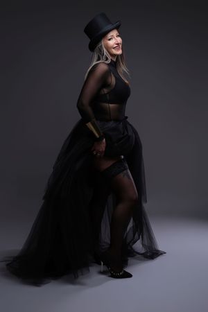 A smiling woman in a black top hat, sheer bodysuit, and flowing black tulle skirt poses elegantly in a studio.