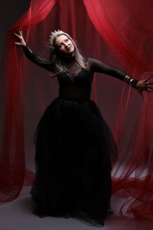 Woman in black ball gown and crown poses dramatically against red flowing curtains in dark fantasy portrait.