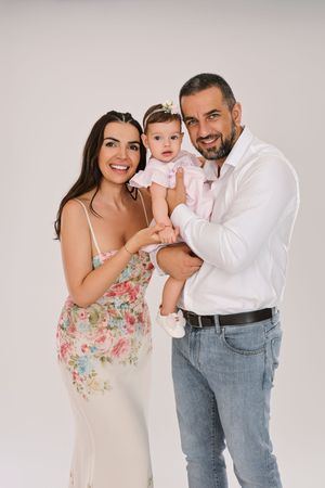 Happy family portrait with smiling parents holding their baby girl in pink dress against white background.