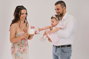 A family of three celebrating a baby girl's first birthday with a white cake in a bright studio setting.