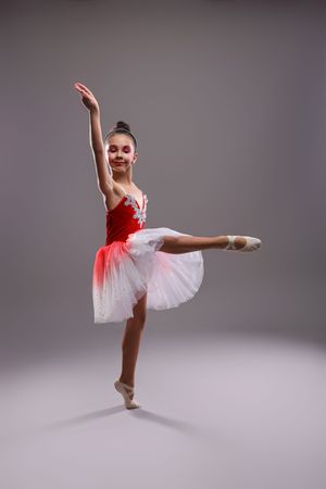 Young girl ballet dancer in red and white tutu performing arabesque pose in ballet slippers on gray background.