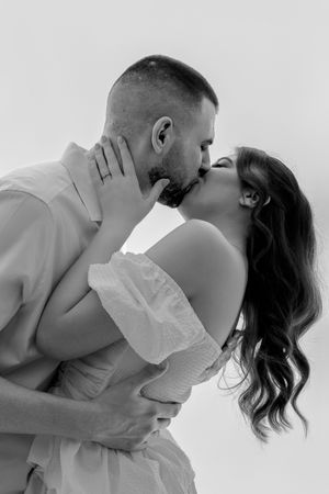 Black and white photo of a romantic couple sharing a passionate kiss, woman in white off-shoulder dress.