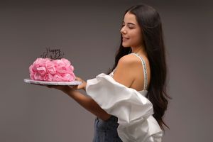 Young woman in white off-shoulder top holding a pink rose birthday cake on a gray background.