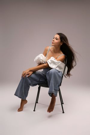 Barefoot woman in white top and wide-leg jeans sits on chair with flowing dark hair against gray background.